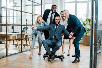 Wall Mural - cheerful businessman sitting on chair with outstretched hands near multicultural coworkers