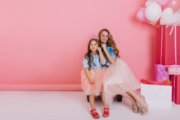 Wall Mural - Graceful mother and daughter in pink skirts and vintage shirts holding carnival masks and laughing at family party. Adorable young woman with trendy hairstyle sitting with little sister next to gifts