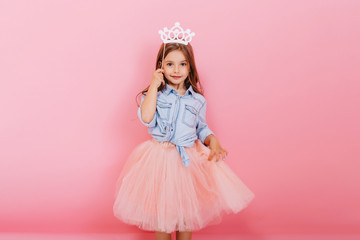 Wall Mural - Cheerful little girl with long brunette hair in tulle skirt holding princess crown on head  isolated on pink background. Celebrating brightful carnival for kids, birthday party, having fun of cute kid