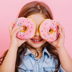 Closeup portrait happy cute little girl in blue shirt holding donuts on eyes, having fun to camera isolated on pink background. Expressing happiness of amazing child, tasty dessert