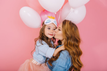 Wall Mural - Attractive curly young woman with party balloons congratulates daughter on birthday and kisses her on the cheek. Little girl in sleep mask with surprised face expression embracing mom at event
