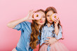 Attractive curly girl in denim shirt embracing little sister and funny posing with delicious donut on pink background. Stylish long-haired mom and cute daughter having fun holding doughnuts as glasses