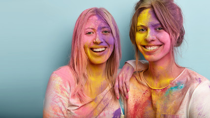 Photo of two sisters stand closely, smile broadly, have happy face smeared with colorful powder, have fun on Holi festival, pose over blue background with copy space aside. Holiday of colors