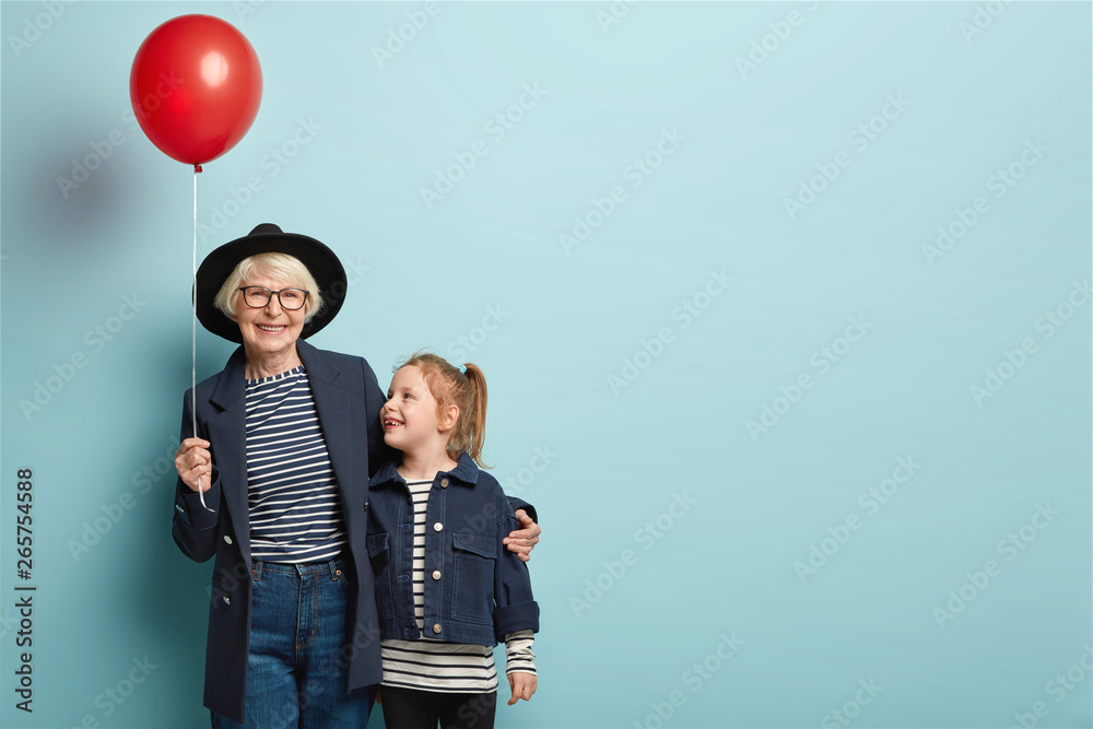 Indoor shot of fashionable senior woman embraces little child, enjoy spending time together, celebrate first day at school, hold air balloon, pose over blue background with blank space on right - obrazy, fototapety, plakaty 