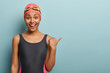 © wayhome.studio  - Slim dark skinned female swimmer in black bathingsuit, points thumb on free space, wears pink swimming cap and goggles, leads healthy lifestyle, poses against blue background. Hobby concept.