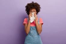Portrait Of Sick African American Woman Sneezes In White Tissue, Suffers From Rhinitis And Running Nose, Has Allergy On Something, Looks Unhealthy, Feels Unwell. Symptoms Of Cold Or Allergy.