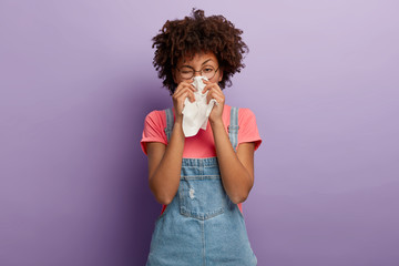portrait of sick african american woman sneezes in white tissue, suffers from rhinitis and running n