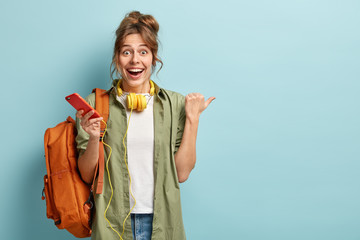 Joyful European girl checks nootification or email box on cellular, enjoys listening favourite music in headphones, has surprised happy reaction on something aside, points thumb at free space