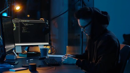 Poster - cybercrime, hacking and technology concept - asian male hacker in hoodie using computer virus program for cyber attack and taking notes in dark room at night