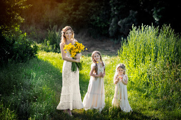 Mothers Day. Beautiful young mother with two daughters on the nature in white dresses and yellow flowers. Mother's love.