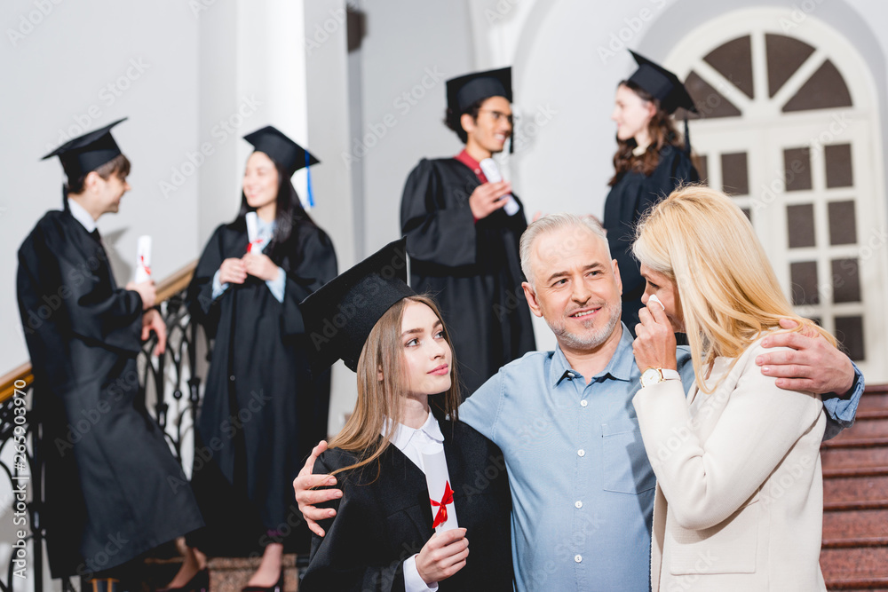 selective focus of attractive girl in graduation cap looking at crying mother while hugging with father near students - obrazy, fototapety, plakaty 