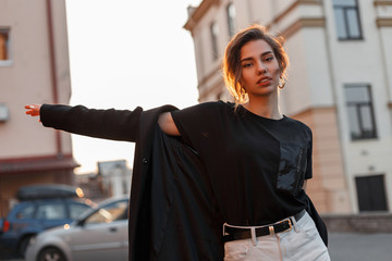 Wall Mural - Attractive beautiful young woman in a stylish T-shirt in a coat in vintage jeans resting outdoors in the city on a background of bright orange sunset. Modern pretty girl model enjoys the sunlight.