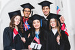 © LIGHTFIELD STUDIOS - selective focus of cheerful students in graduation gowns holding flags of different countries and taking selfie on smartphone