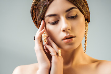 Wall Mural - young nude woman with hands near face, shiny makeup, golden rings and earrings in turban isolated on grey