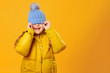 © Olga - Closeup portrait of a cheerful little girl in jacket over yellow background. The child hides his eyes under the cap.