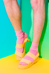 Wall Mural - Male hairy legs in socks staying in women's sandals on bold background in the corner with strong shadows. Minimal art. Pride concept. Body part