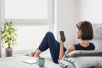 Wall Mural - Young beautiful woman resting at home using smart phone. Happy girl texting on her mobile phone online. Relaxation, freelance work concept