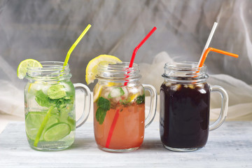 cold beverages mojito lemonade with mint and lemon, strawberry lemonade, coffee cold brew drink with ice and two tubules on a white wooden table top in glass mug jar with handle on rustic background