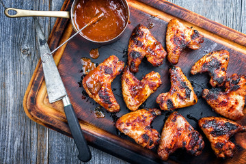 Wall Mural - Traditional barbecue chicken wings with hot chili sauce as top view on a burnt cutting board
