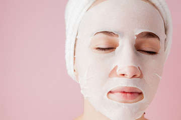 Wall Mural - Beautiful young woman is applying a cosmetic tissue mask on a face on a pink background. Healthcare and beauty treatment and technology concept