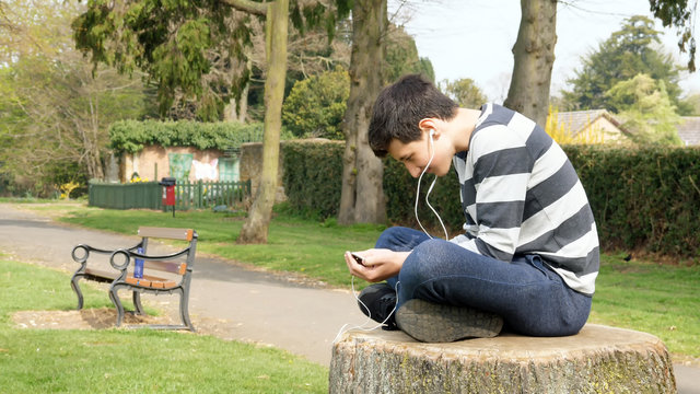 teen boy with smart phone listening or talking while sitting on stump in british park. teenager and social media concept