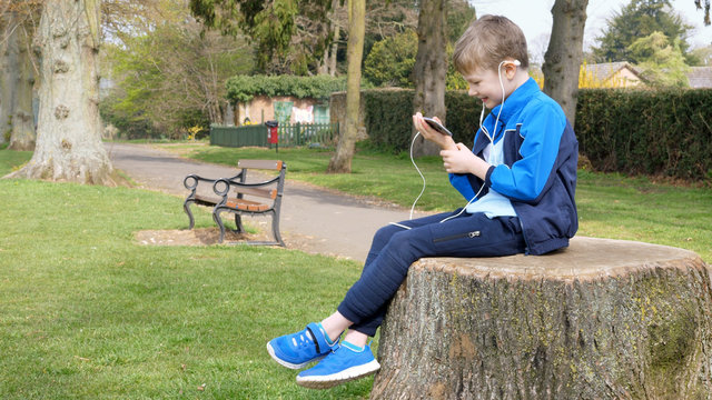 teen boy with smart phone listening or talking while sitting on stump in british park. teenager and social media concept