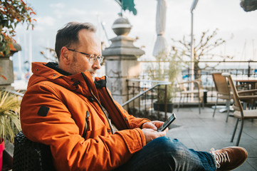 Outdoor portrait of middle age man wearing eyeglasses and orange winter jacket, holding smartphone