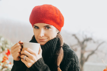 Close up portrait of young woman drinking hot tea on the balcony on a cold foggy daym wearing warm pullover and red beret