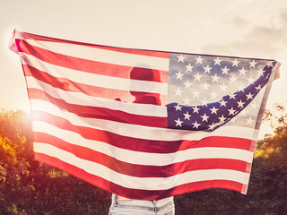 Handsome man waving an American flag against a background of trees and blue sky. View from the back, close-up. National holiday concept