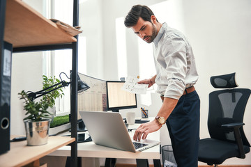 Fototapete - Multitasking. Side view of successful bearded trader or businessman in formal wear working with laptop while standing at his modern office.
