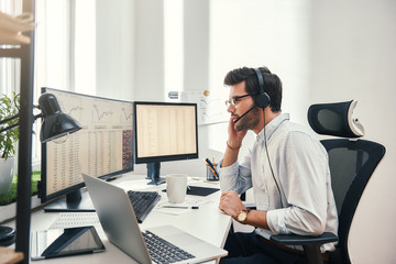 Fototapete - Always available. Side view of young bearded trader in headset talking with client and looking at monitor screen with trading charts and financial data while sitting in his modern office.