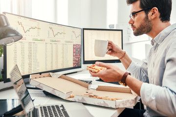 Food for productive work. Side view of young bearded trader in eyewear is eating hot pizza while looking at monitor screen with trading charts and financial data in his modern office.