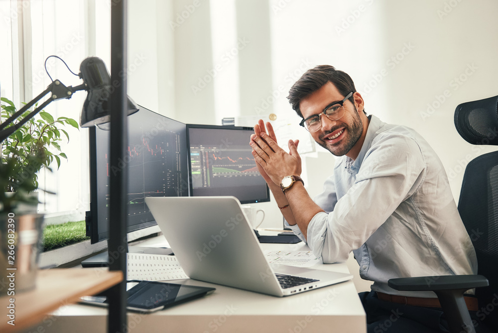 Young and successful. Happy bearded trader in formal wear and eyeglasses looking at camera and smiling while sitting in his modern office. - obrazy, fototapety, plakaty 