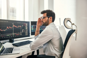 Fototapete - Full concentration. Young and smart businessman in formal wear is adjusting his eyeglasses and using laptop while while sitting in his office in front of computer screens with trading charts
