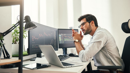 Fototapete - Trading strategy. Successful young trader in eyeglasses looking at analyzing trading charts on computer screens in while sitting in his modern office.