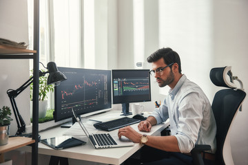 Fototapete - Busy working day. Young bearded trader in eyeglasses working with laptop while sitting in his modern office in front of computer screens with trading charts.