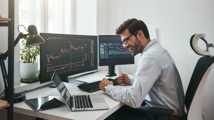 Poster - Lucky day. Happy young businessman or trader in formalwear and eyeglasses using laptop and smiling while sitting in his modern office