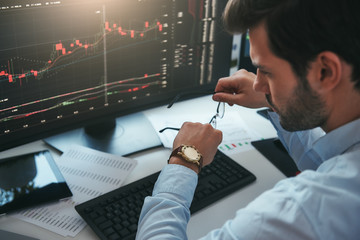 Fototapete - Time to trade. Bearded male trader looking at watch on his hand while working with data and charts on computer screens in his modern office.