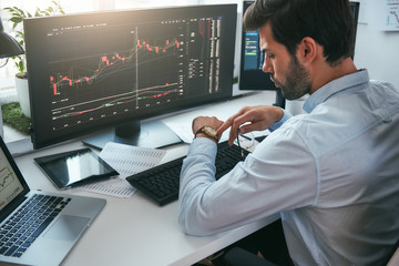 Fototapete - What time is it? Back view of bearded male stock market broker looking at watch on his hand while working with data and charts on computer screens in his modern office.