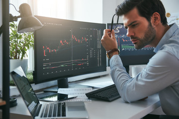 Fototapete - Successful investor. Serious young male trader holding eyeglasses and analyzing data and graphs on his laptop while working in his modern office
