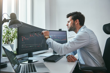 Fototapete - Analyzing data. Thoughtful young trader is pointing at graphs on computer screen while working in his modern office.