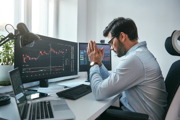 Fototapete - Hoping for the success. Side view of tired young businessman or trader holding palms together while working with data and charts on computers at his modern office.