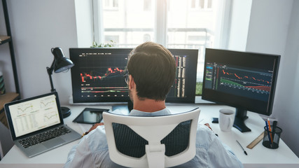 Fototapete - Trading stocks online. Back view of young businessman or trader working with graph and charts on computers at his modern office