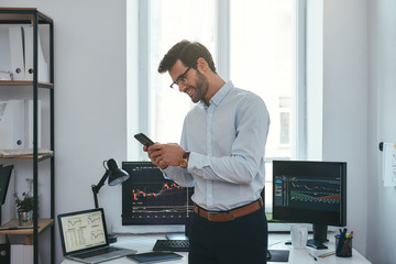 Fototapete - Modern business. Happy and successful trader in formal wear is looking at trading graphs and reports on his smartphone for investment purpose and smiling while standing at his office