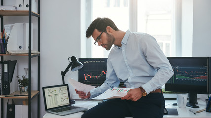 Fototapete - Busy working day. Young successful trader or businessman in formal wear and eyeglasses sitting at modern office in front of computer screens with charts and reading market report