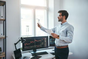Fototapete - Trading strategy. Smart and young trader in eyeglasses looking at financial reports and analyzing trading charts while standing in front of computer screens in modern office.