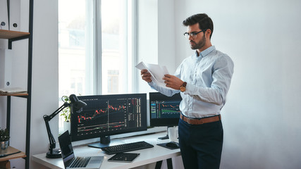 Fototapete - Analyzing data. Thoughtful trader in eyeglasses holding a financial report and analyzing trading charts while standing in front of computer screens in modern office