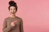 Fototapeta  - Portrait of happy amazed young beautiful lady with curly dark hair, heard the cool news, broadly smiling, looking at the camera, pointing with finger to copy space isolated over pink background.