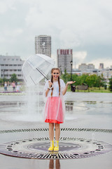 Young wet pretty girl with two braids in yellow boots and with transparent umbrella stands inside of fountain.