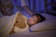 © DN6 - Mother putting blanket on and stroking head for her little girl daughter on bed in a dark bedroom at night, Child asian girl hug teddy bear, Comfortable children at home concept